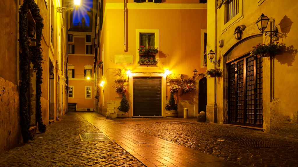 An ochre-walled Roman lane at night lit by a single street lamp