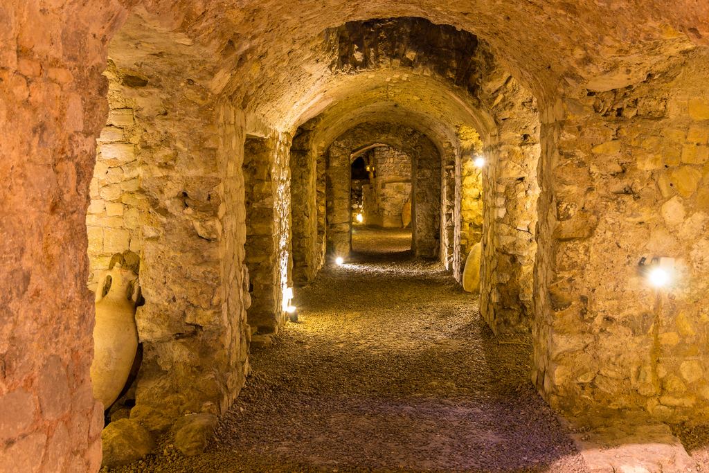 A long barrel-vaulted Roman storage corridor below street level, lit along its length, with an amphora set in a niche