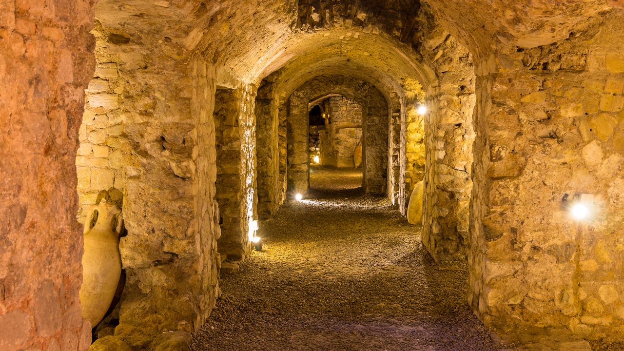 A long barrel-vaulted Roman storage corridor below street level, lit along its length, with an amphora set in a niche