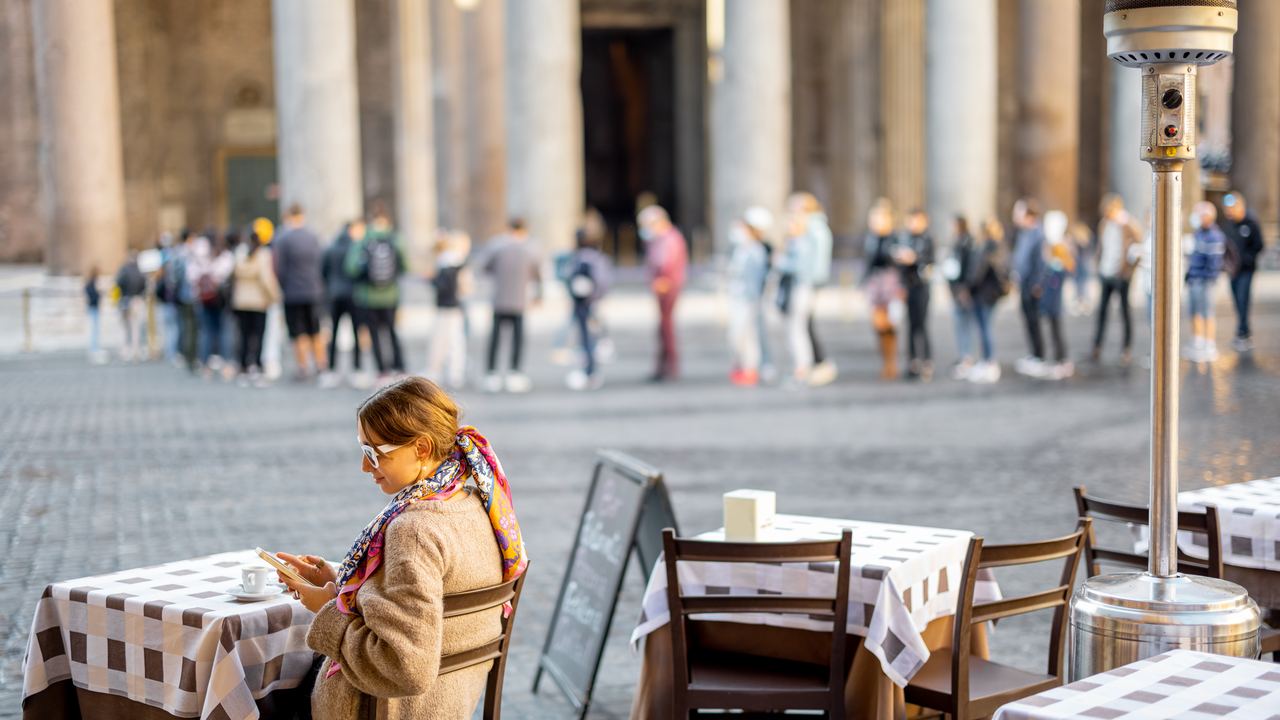 A woman at a cafe table in Piazza della Rotonda with the line of visitors and the Pantheon's columns behind her