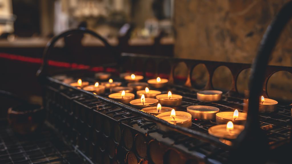 Rows of small votive candles burning on a metal stand inside a Catholic church