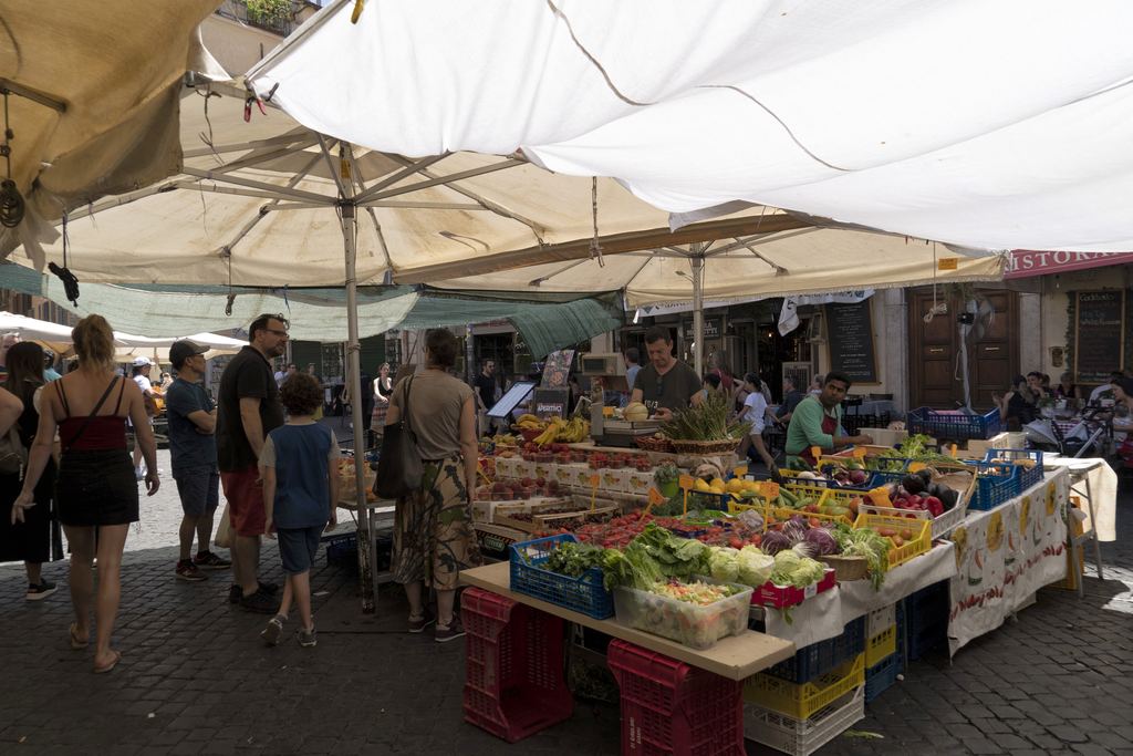 Fruit and vegetable stalls under white awnings in Campo de' Fiori, shoppers crossing the cobbles