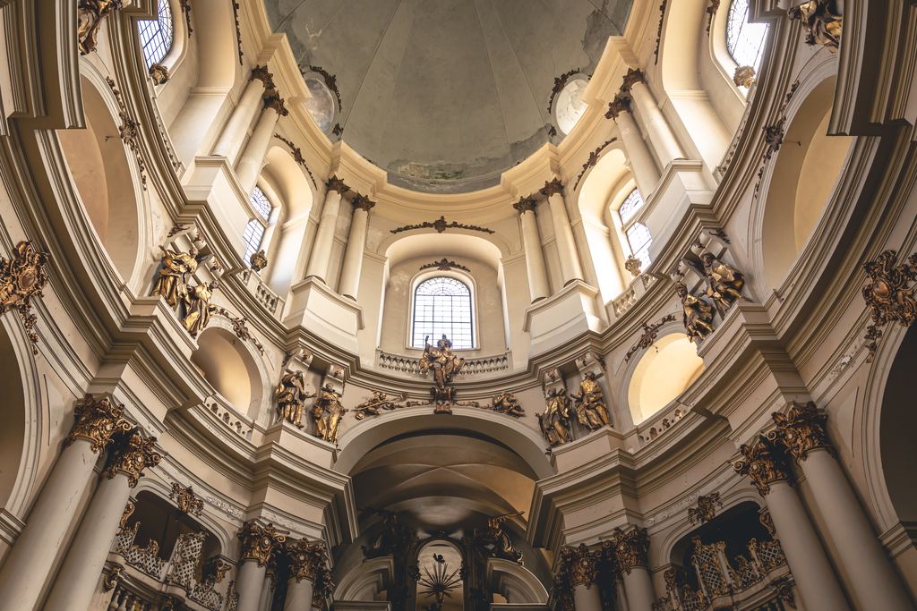 The inside of a baroque church looking up: cream stucco, gilded ribs, statues in niches and tall arched windows