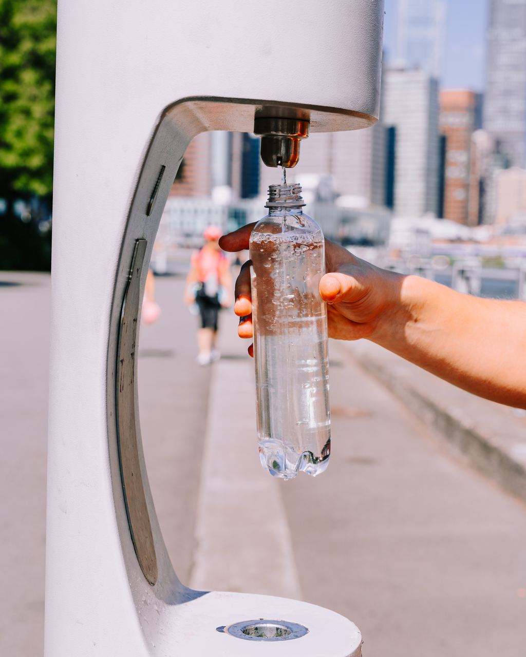 A hand holding a reusable bottle under the spout of a public drinking fountain in a city street