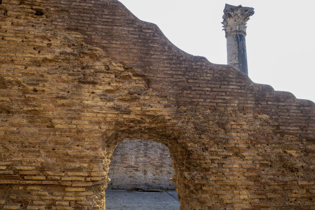 A Roman brick arch and wall standing at Ostia Antica, a marble Corinthian capital visible beyond it