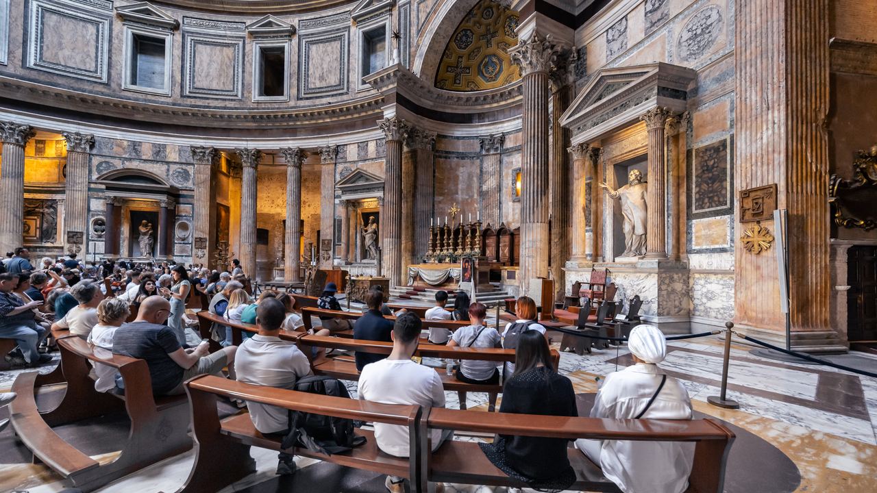 Visitors seated on benches facing the high altar inside the Pantheon in Rome, with the apse and the coffered drum behind