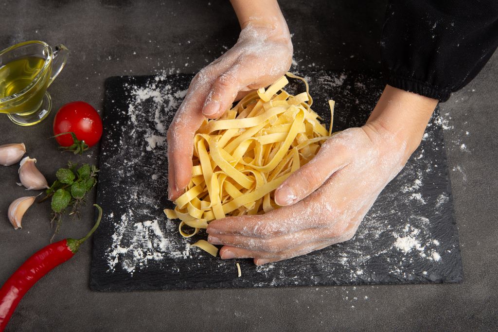 Floury hands lifting a nest of fresh tagliatelle from a dark board, with oil, tomatoes, garlic and basil alongside