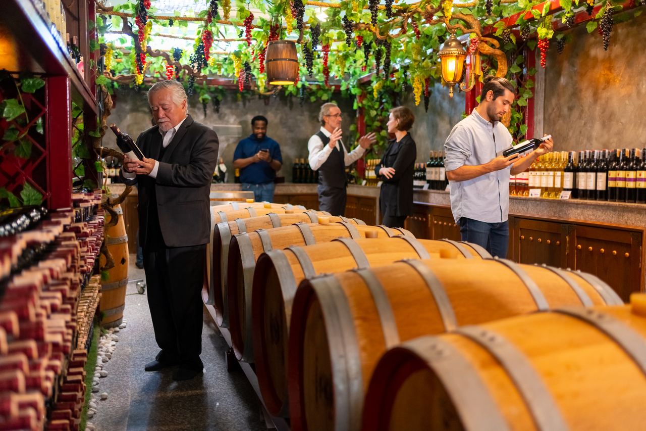 A small group standing with glasses in a cellar lined with wooden barrels and shelves of bottles