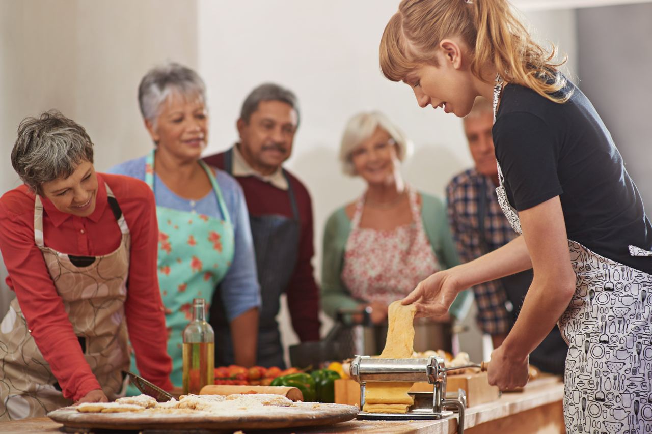 A cooking class gathered round a table while one person feeds pasta dough through a hand-cranked roller