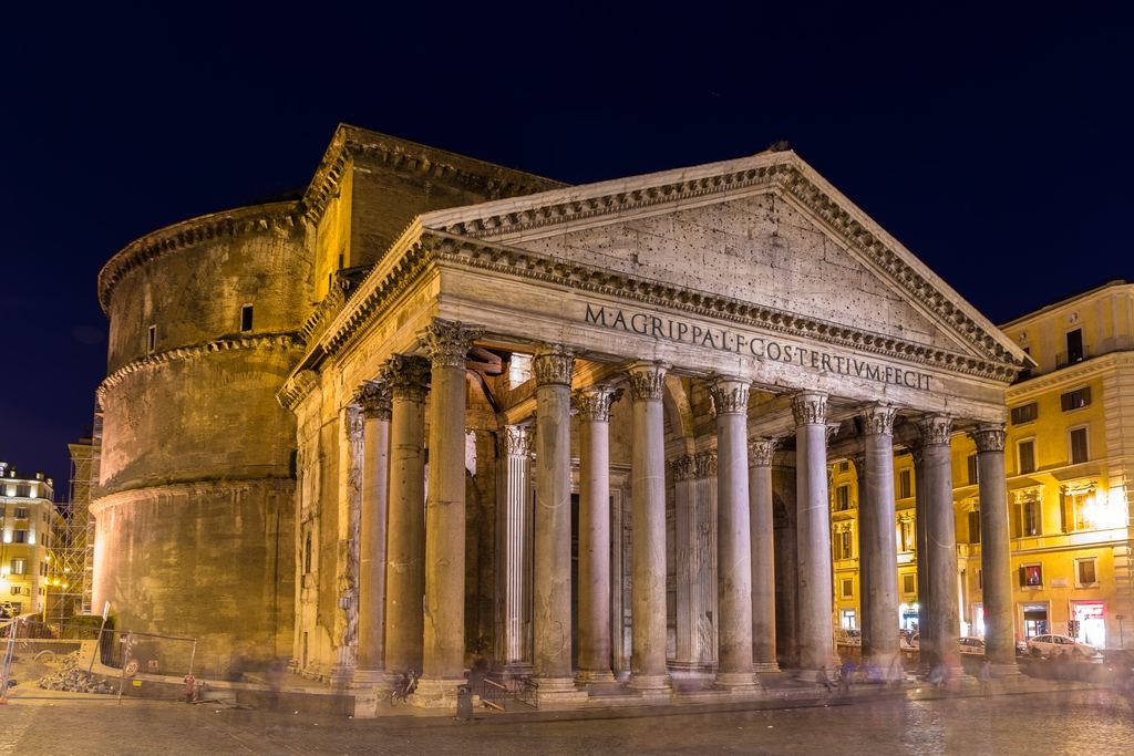The Pantheon lit amber at night from an angle, with people and bar signs in the square below