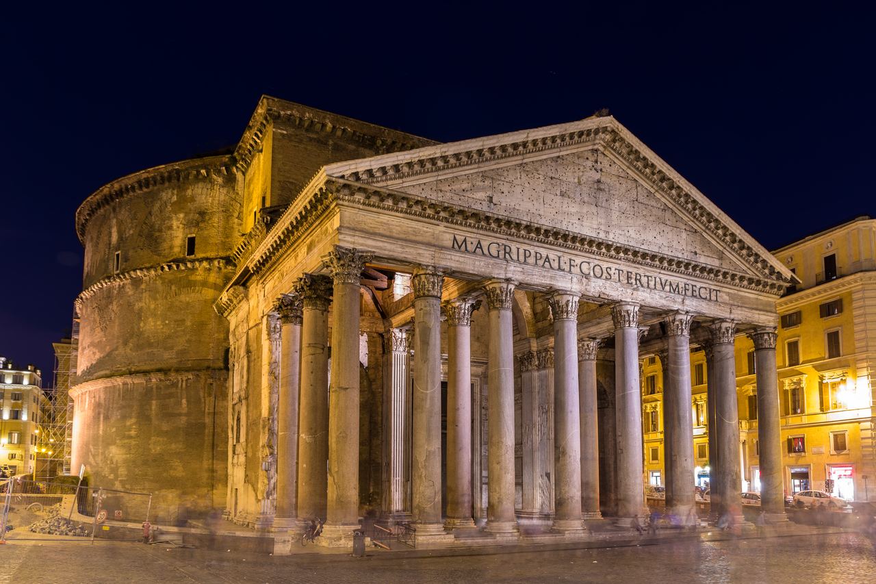The Pantheon lit amber at night from an angle, with people and bar signs in the square below